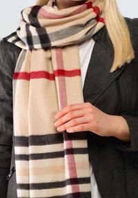 Femme portant une écharpe à carreaux beige, noir et rouge sur une chemise blanche et une veste noire, tenant le bord de l'écharpe de la main droite.