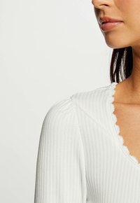 Primo piano della parte inferiore del volto di una donna, collo e spalla, che indossa un top bianco a coste con scollatura ondulata e maniche lunghe.