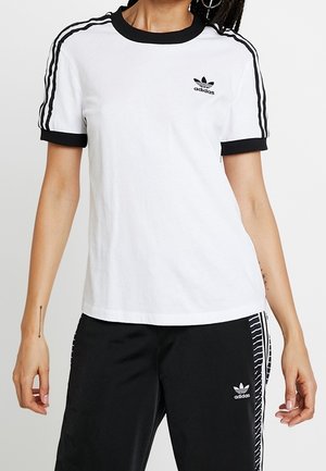 Personne portant un t-shirt Adidas blanc avec col et manches noirs, pantalon Adidas noir avec des bandes blanches et des logos sur les deux vêtements.