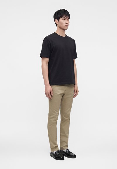 Jeune homme debout face à l'avant, portant un t-shirt noir à manches courtes, un pantalon beige et des chaussures habillées noires sur un fond blanc uni.