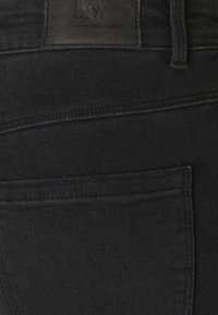 Jeans din denim negru, cu o etichetă inscripționată "JOY" și "THE DENIM STORY", având un buzunar pe spate și o textură netedă.