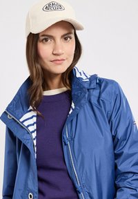 Donna con capelli castani che indossa un cappello bianco, una giacca blu con rifiniture a righe e un maglione viola, guarda dritto davanti a sé con un'espressione neutra.