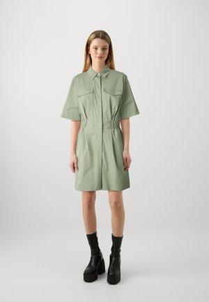 Rochie verde cu guler, mâneci scurte, buzunare frontale și talie elastică. Pur și simplu cu cizme negre cu platformă pe un fundal deschis.