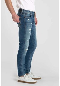 Vaqueros de mezclilla azul con un corte slim, que presentan parches desgastados y un diseño clásico de cinco bolsillos. Combinados con zapatillas blancas.