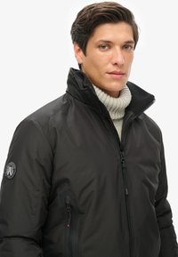 Chaqueta aislante negra con un alto cuello, suéter gris de canalé debajo, bolsillo con cremallera lateral y un logo en el brazo izquierdo.
