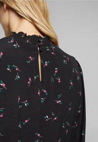 Schwarze Bluse mit floralem Druck in Pink und Grün, mit einem Schlüsselrücken und einem rüschengeschmückten Ausschnitt. Weicher, leichter Stoff.