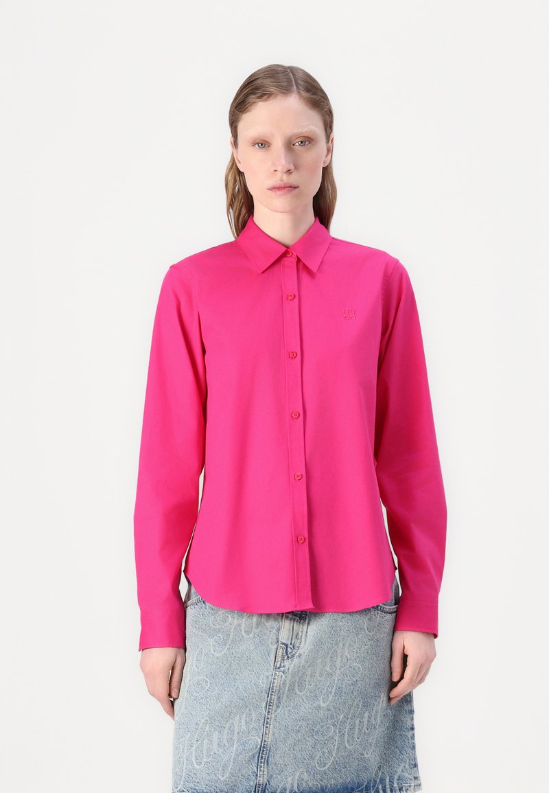 Fuchsia long-sleeve shirt with a collar, button closure, and small logo detail. Worn over light denim skirt with repeating text pattern.