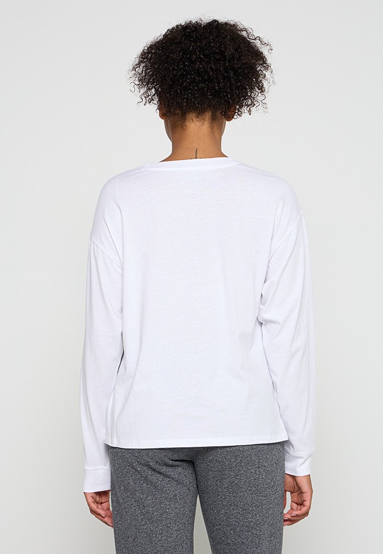 Person mit lockigem Haar, der ein weißes langärmliges Shirt und graue Hosen trägt, von hinten vor einem einfarbigen hellen Hintergrund gezeigt.