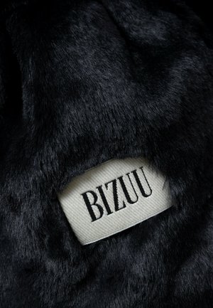 Czarna tekstura sztucznego futra z tkaną metką z napisem "BIZUU" w czarnych literach na kremowym tle. Materiał wydaje się miękki i puszysty.
