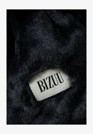 Czarna tekstura sztucznego futra z tkaną metką z napisem "BIZUU" w czarnych literach na kremowym tle. Materiał wydaje się miękki i puszysty.