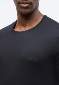 Άνδρας που φοράει μαύρο μπλουζάκι BOSS με λαιμόκοψη στρόγγυλη, εμφανιζόμενος από τον λαιμό έως το πάνω μέρος του στήθους, με απλό φόντο.