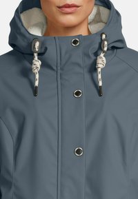 Close-up van een persoon die een grijze hoodie-jack draagt met witte fleece voering, toggle trekkoorden en zwarte drukknoopjes aan de voorkant.