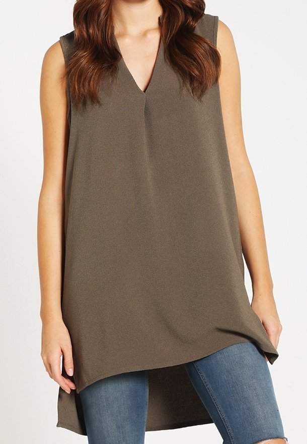 Femme portant une tunique marron sans manches avec col en V et ourlet asymétrique sur un jean bleu déchiré sur fond blanc.