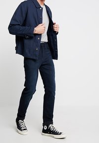 Tmavě modrá bunda s límečkem, šedé tričko, tmavé džíny a černé vysoké tenisky s bílou podrážkou a černými tkaničkami.