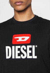 Svart bomull t-shirt med en framträdande röd rektangulär logotyp med vit text "DIESEL" och ett stiliserat "D" emblem ovanför.