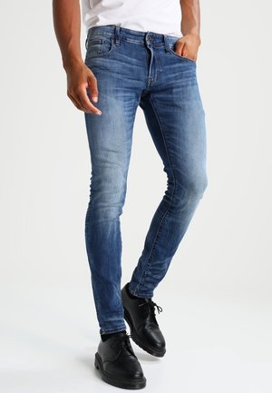 Homme portant un jean slim bleu délavé, des chaussures habillées noires et une chemise blanche, une main dans la poche, debout devant un fond blanc.