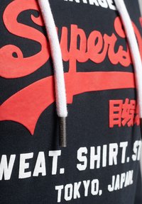 Gros plan d'un sweat à capuche noir avec le texte rouge "Super", des cordons blancs et des lettres blanches en bloc indiquant "SWEAT. SHIRT. TOKYO, JAPAN."