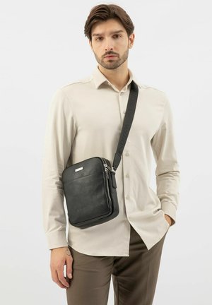 Borsa a tracolla in pelle nera con superficie testurizzata, dettaglio della cerniera argentata e tracolla regolabile, indossata da una modella con una camicia chiara.