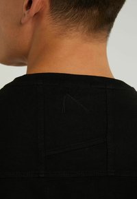 T-shirt noir avec un col rond, présentant un petit détail de couture inachevée à l'arrière et un tissu lisse et texturé.