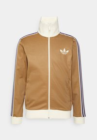 Brun zip-up jacka med hög krage, vita accenter, två sidofickor och lila-randiga detaljer på ärmarna. Adidas-logotyp på vänster bröst.