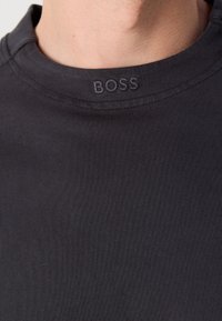 Gros plan d'un t-shirt noir à col rond avec broderie en relief du logo BOSS sur le col, porté par une personne.
