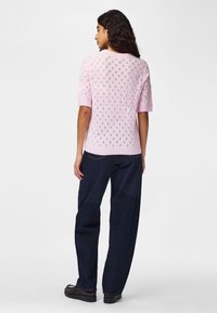 Femme aux cheveux bouclés portant un pull en tricot à manches courtes rose clair et un jean ample bleu foncé, debout de dos.