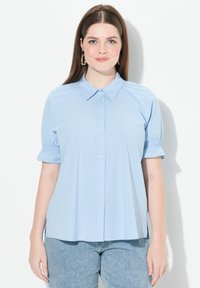 Camicia azzurra chiara con colletto, maniche corte a sbuffo con polsini elastici e orlo dritto. Tessuto morbido.
