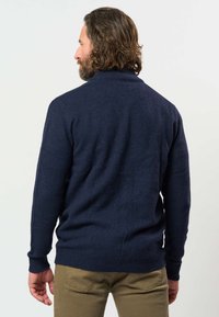 Man met kastanjebruin schouderlang haar en baard, draagt een donkerblauwe trui en kaki broek, met zijn rug naar een effen achtergrond.