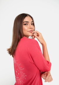 Rožinė per didelė t-shirt su trumpomis rankovėmis, su balta saulės ir mėnulio grafika nugaroje, demonstruojanti minkštą medžiagą ir laisvą siluetą.