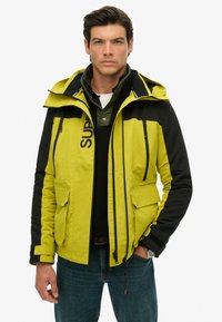 Yellow and black waterproof jacket with a hood, zippered pockets, and logo detail. Features a textured lining and elastic cuffs.