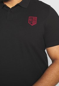 Detailní záběr na osobu v černém polokošili s malou červenou vyšitou nášivkou na levé straně hrudi.