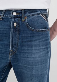 Jeans in denim blu con una leggera elasticità, dotati di cinque tasche, minuteria metallica e cuciture a contrasto. L'etichetta mostra il nome del marchio sulla vita.