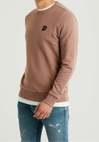 Bruine sweatshirt met een ronde hals en geribbelde manchetten, met een klein zwart logo op de borst. Draagt over een wit ondershirt.