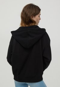 Felpa nera oversize con tasca a marsupio e polsini a costine. Caratterizzata da un cappuccio con cordino, una texture morbida e un fit rilassato.