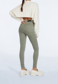 Jeans skinny verde oliva con bolsillos traseros y cintura alta, combinados con un suéter de punto color crema con un diseño corto.