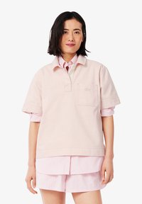 Helles rosa Poloshirt mit einer Fronttasche, kurzen Ärmeln und einem Kragen, über einem pink-weißen gestreiften Hemd mit Knöpfen getragen.