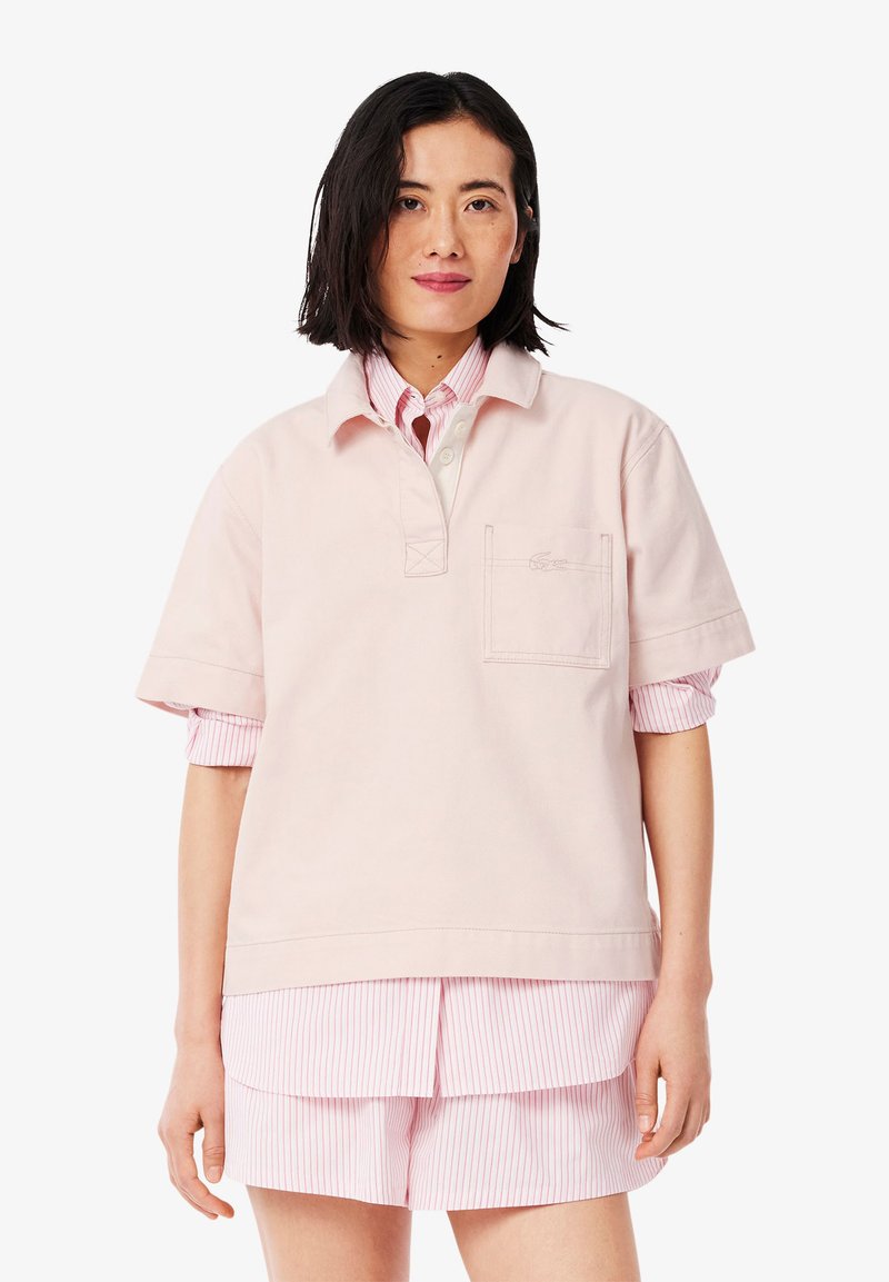 Helles rosa Poloshirt mit einer Fronttasche, kurzen Ärmeln und einem Kragen, über einem pink-weißen gestreiften Hemd mit Knöpfen getragen.