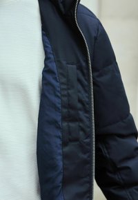 Jachetă căptușită de culoare bleumarin cu fermoar frontal, material texturat pe corp și mâneci netede. Prezintă o buzunar cu clapă pe partea stângă.