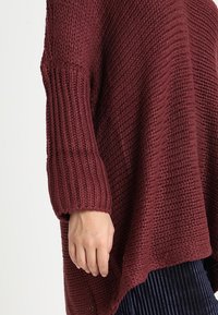 Burgundy stickad tröja med strukturerad mönster, ribbade ärmar och lös passform, med en hand synlig nära fållen.