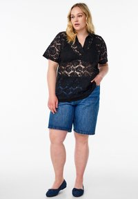Blusa nera in pizzo con maniche corte e colletto, abbinata a pantaloni corti in denim blu. Il modello indossa scarpe basse blu, in piedi con una mano in tasca.
