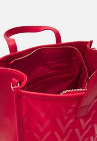 Röd handväska med en slät läderutsida, som har guldtonade dragkedjor och ett foder inuti av tyg. Chevronmönster synligt.
