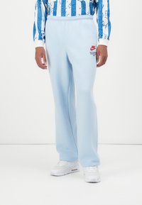 Gaiši zilas sporta bikses, kas izgatavotas no mīksta auduma, ar elastīgu jostu un mazu sarkanu Nike logotipu uz kreisā augšstilba.