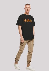 Schwarzes Baumwoll-T-Shirt mit einem auffälligen orangefarbenen und gelben "Def Leppard"-Logo. Kombiniert mit beigen Cargo-Hosen und weißen Turnschuhen.