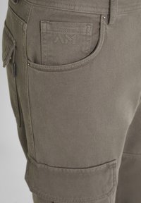 Primo piano di pantaloni cargo brown che mostrano la tasca anteriore, una piccola tasca rettangolare cucita e una tasca laterale con pattina con cuciture visibili.