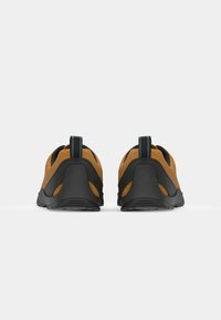 Brown suede shoes with a black rubber sole, featuring cut-out accents and a blue pull tab at the back. Minimalistic design.