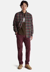 Kostkovaná flanelová košile v burgundské a zelené barvě, navrstvená přes hnědé tričko s potiskem. Burgundské manšestrové kalhoty a béžové boty doplňují outfit.