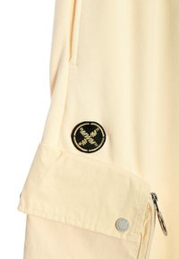 Chaqueta de tejido beige claro con un bolsillo con cremallera, logotipo redondo bordado y una textura suave. El diseño incluye un detalle de manga doblada.