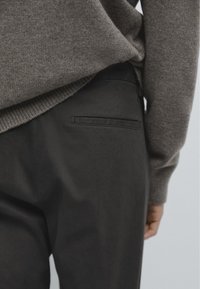 Gros plan sur une personne portant un pull en tricot marron et un pantalon noir sur mesure avec une poche passepoilée unique à l'arrière.
