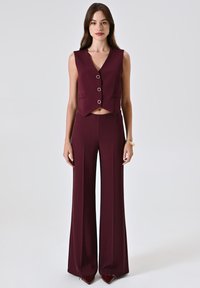 Completo bordeaux a due pezzi con gilet senza maniche, caratterizzato da un scollo a V e tre bottoni, abbinato a pantaloni a gamba larga a vita alta.