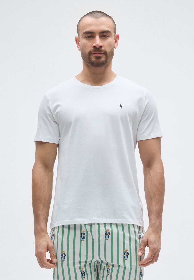 Fehér pamut póló rövid ujjakkal és kerek nyakkivágással, amelyen kis tengerészkék logó található a mellkasnál. Zöld csíkos nadrággal viselve.